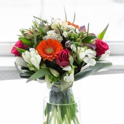 This captivating bouquet, hand-crafted by Abbey Wood Florist, seamlessly blends colour, form, and feeling into an unforgettable arrangement. Creamy peach gerberas form a luminous ring around vibrant orange centers, their symmetry reminiscent of the balanced paths winding through Lesnes Abbey Woods. Deep, luxurious fuchsia roses nestle amidst delicate white alstroemeria and filler blooms, providing a harmonious contrast that draws attention to each unique petal. The base is hugged by broad, shiny aspidistra leaves, their protective arc reminiscent of the ancient walls and lush greenery of Abbey Wood, while slender pointed leaves add a lively, dynamic energy. Blush pink and soft white blossoms peek from among the greens, enhancing the bouquet's rich, layered effect-much like the changing light over the local woodlands. Displayed in a sleek glass vase, with neatly arranged stems visible, the entire ensemble glows softly under diffuse daylight, evoking a morning stroll through woodland trails. Gentle notes of rose and lily glide on the air, fresh and comforting. Perfect for marking a special occasion-a birthday, anniversary, or welcome to a new neighbour-this bouquet embodies optimism, gratitude, and the heartfelt care that Abbey Wood Florist pours into every floral creation for the local community.